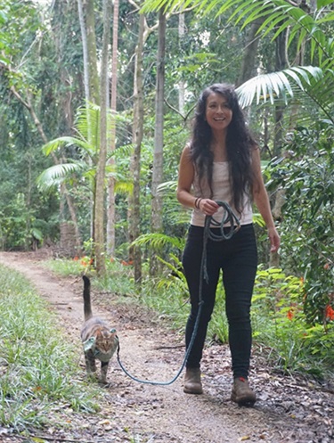 Image of a woman with long dark hair walking along a rainforest path with her cat on a lead.