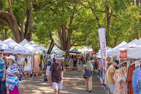people looking at market stalls shaded by large trees