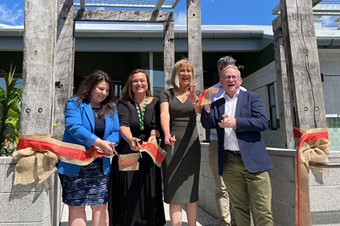 Three women and a man cutting red ribbon tied between two wooden posts