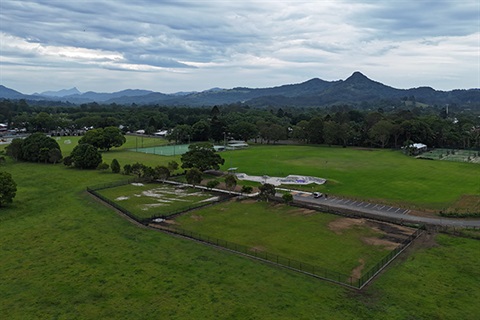 High view of two fenced, grass paddocks with skatepark, sportsfield and mountains in background