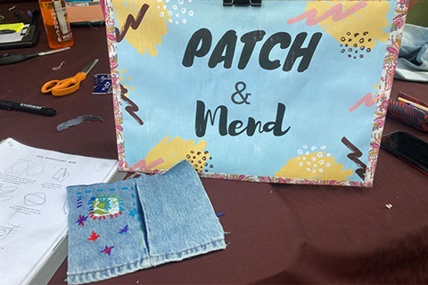 piece of denim fabic with colourful needlework next to a sign saying patch and mend